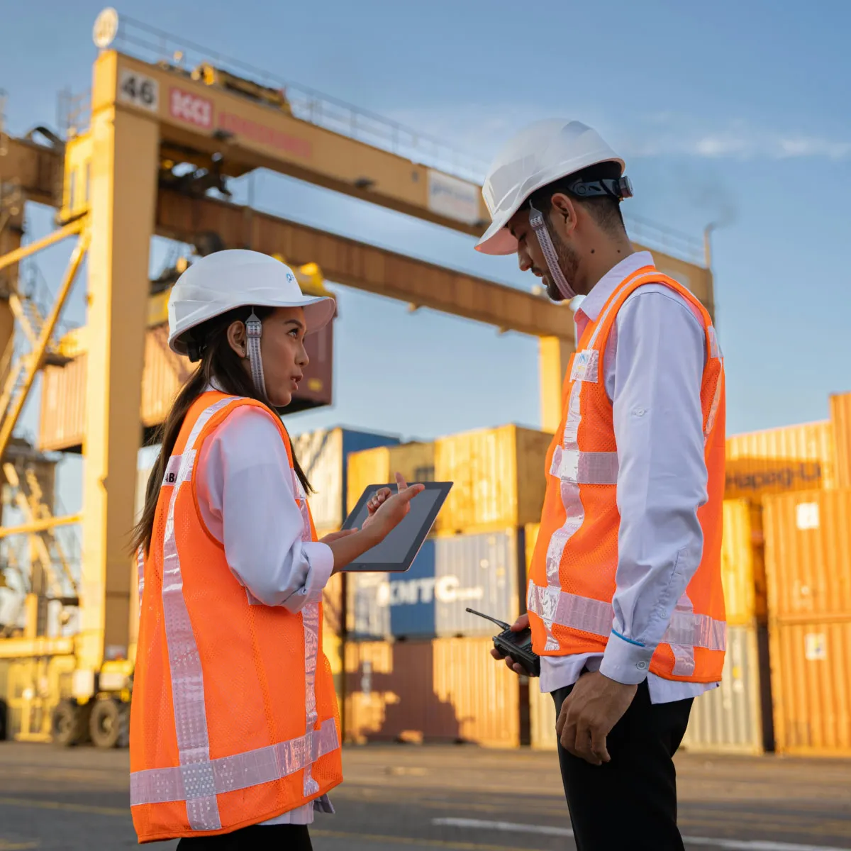 Two construction workers wearing white helmets and orange safety vests discussing with a tablet and a walkie-talkie at a shipping container yard.