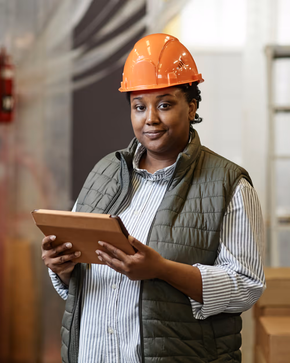 Woman wearing an orange hard hat and green vest holding a tablet in an industrial setting.
